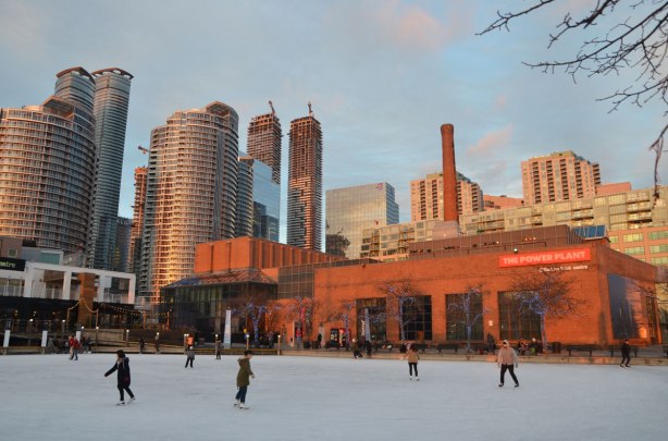 late afternoon with the sun low in the sky casting yellowish glow on the world, Toronto skyline in the background with its condos and construction cranes, also The Power Plant Center and art gallery. In the foreground is the frozen Natrel Pond of Harbourfront and on it people are skating. 