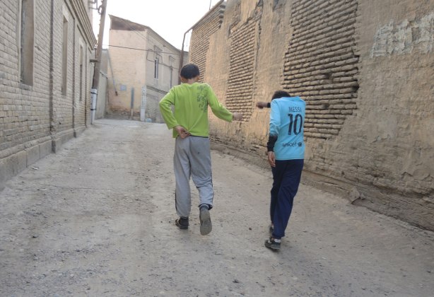 two boys laugh as they walk away, walking down a street with a tall stone wall on one side, and old houses on the other 