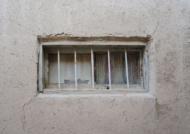 a wall made of mud and straw, side of a house, with an old window in the house. The window has bars on the outside, curtains on the inside, Bukhara, Jewish quarter, 