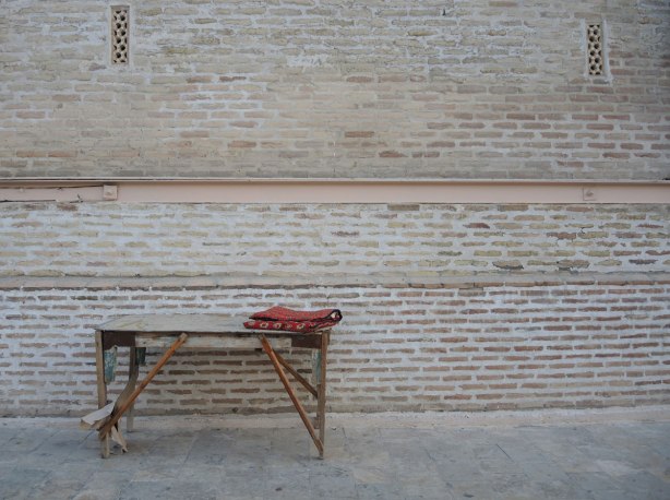 an old brick wall, an old wood bench, and a folded red carpet at one end of the bench. 