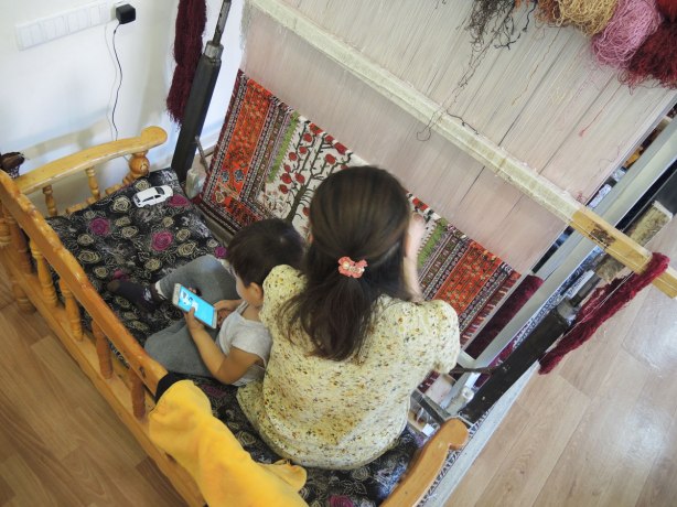 a woman is working on a loom, making a carpet, a young boy sits with her playing games on a smartphone 