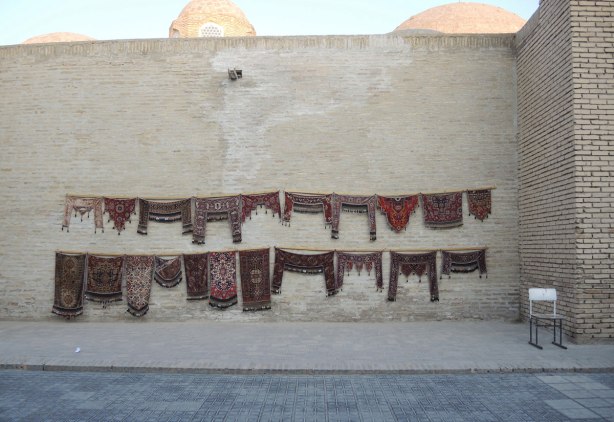 small carpets for sale, strung up against on old brick wall outside, the carpets are designed to fit over the top of a doorway, mostly in dark red and browns, 
