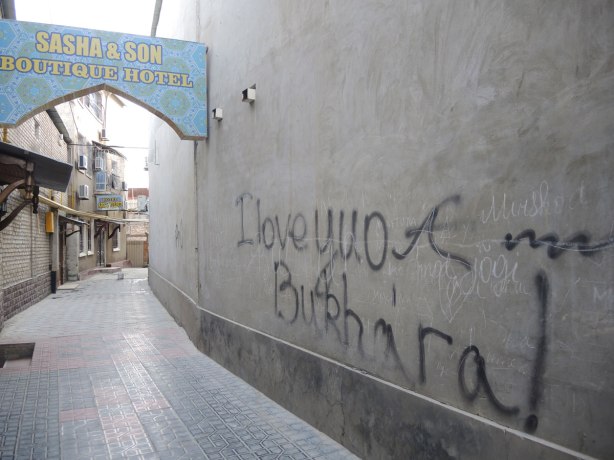 graffiti on a wall in the Jewish quarter of Bukhara, in large black letters someone has written I love you Bukhara. Across the alley is the hotel, Sasha and sons. 