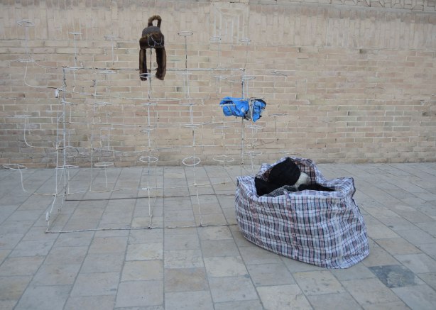a wire rack is empty except for one hat, a full bag sits on the ground beside the rack. An old brick wall is behind. The remains of an outdoor hat seller who has closed up shop for the day 