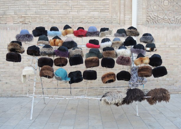 Against an outdoor wall, many fur hats , some are dyed shades of blue or red, for sale in Bukhara Uzbekistan