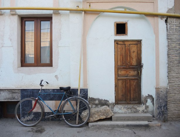 an old house, greyish white, with a wood frame window and old wood door. A bike is parked outside the door. Stone step, yellow pipe for gas runs above the doorway 