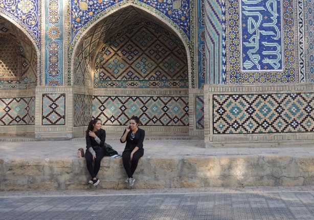 two young woman sitting outside a madrassah. One is sticking her tongue out as she talks on her phone, the other is laughing. 
