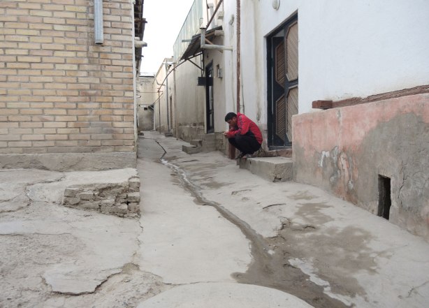 a man in a red sweatshirt sits on a front step, looking at his phone, in an alley with old buildings. 