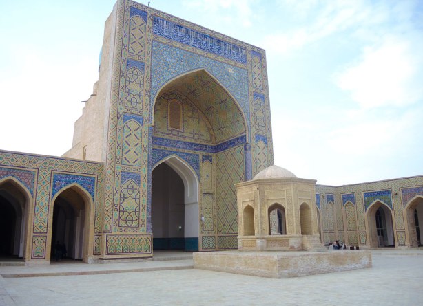 mosque kalyan in bukhara, with its large brick facade over the pointed arch entrance, lots of blue tile, with a small well in front with a brick building like structure over it. 