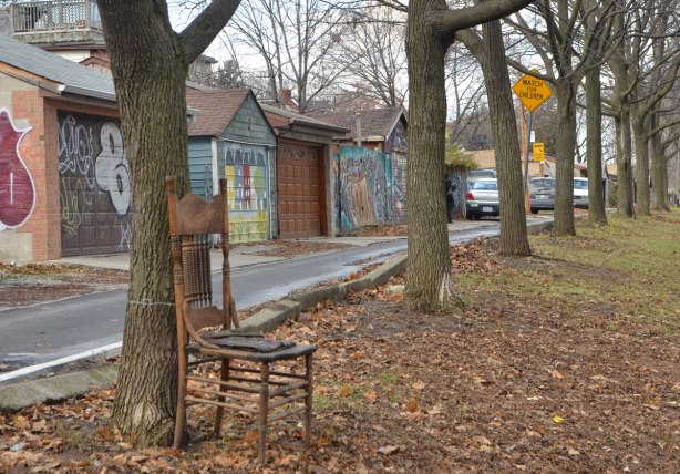 a wooden chair is tied to a tree, autumn leaves on the ground, a row of garages in the background, a number of large trees, 