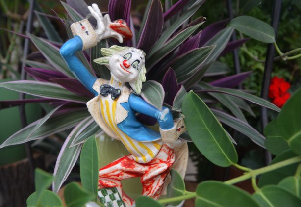 A small figurine of a clown holding his hat above his head, blue jacket, red pants, black bowtie, small figurine in front of a plant with green and purple leaves. 