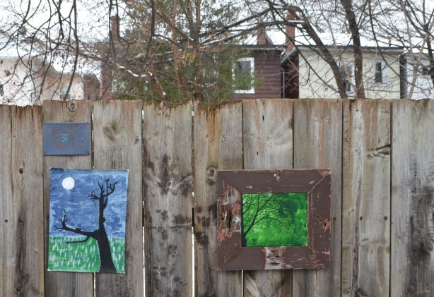 small amateur paintings displayed on a wood fence, with trees and houses in the background, snowy day, Craven Road, trees on blue and green background