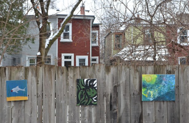 small amateur paintings displayed on a wood fence, with trees and houses in the background, snowy day, three paintings, one is a fish