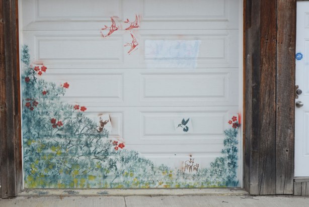 stencil art on a white garage door. greenery, a hummingbird, three swallows in flight, a deer, some red flowers 