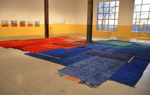 main exhibit room of The Power Plant Contemporary Art Gallery, large room with two large windows in which the low afternoon sun is shining. Exhibit by Yto Barrada consisting of red, green and blue carpets on the floor. some framed pictures on the far wall, walls have been painted yellow up to about 4 feet from the floor. 