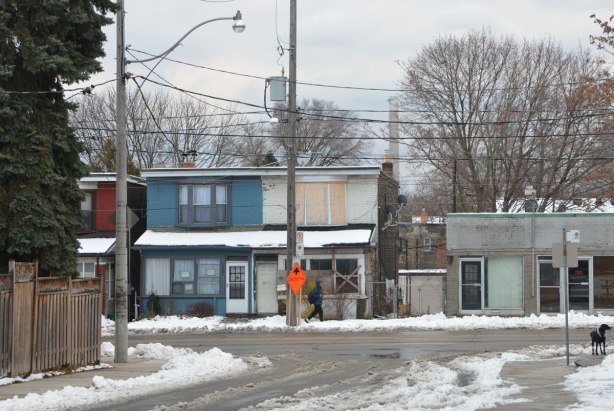 looking towards a street with two sem-detached houses, one is boarded up and the other isn't. A construction sign, a pedestrian on the sidewalk, a black dog. 