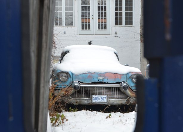 looking through a hole in a blue fence, an old light blue car is parked in a backyard, covered with snow, the white of the house is behind the car. 