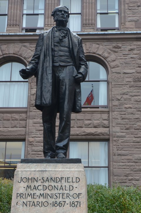 statue of a man, John Sandfield Macdonald, in front of the parliament buildings at Queens Park. An Ontario flag is reflected in the windows of the building. 
