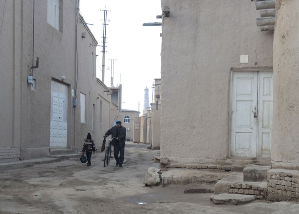 a man walks his bike while a gilr walks beside him carrying her scholl bags, morning, in an alley in khiva