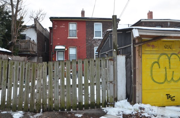 Back of a house in a lane. Gate covering bac is lopsidded, on an angle. The garage that faces the alley has a bright yellow door. Two storey semi detached houses, one brown and one reddish. 