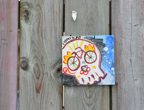 a small painting that has been hung a fence. A white skull with the eyes drawn as the wheels of a bike. The words say I love my bike, with the word love being a small heart. 