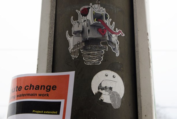 two stickers on a pole. The top one is a lovebot grominator joint venture hybrid character with a very long red tongue. The other is a little round faced guy with his tongue sticking out.
