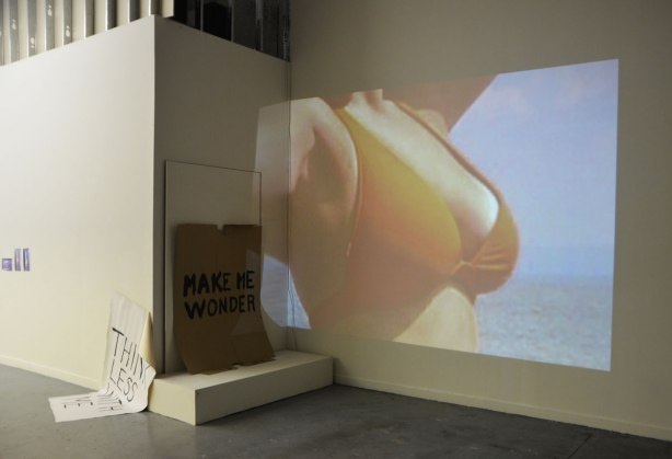 walls of a n art gallery with some of the work of Oscar Figueroa. A projection of a woman's chest in a bikini top, a piece of brown cardboard with the words 'Make me wonder' and a piece of paper, large white paper, half on the floor and half propped up on the wall that says 