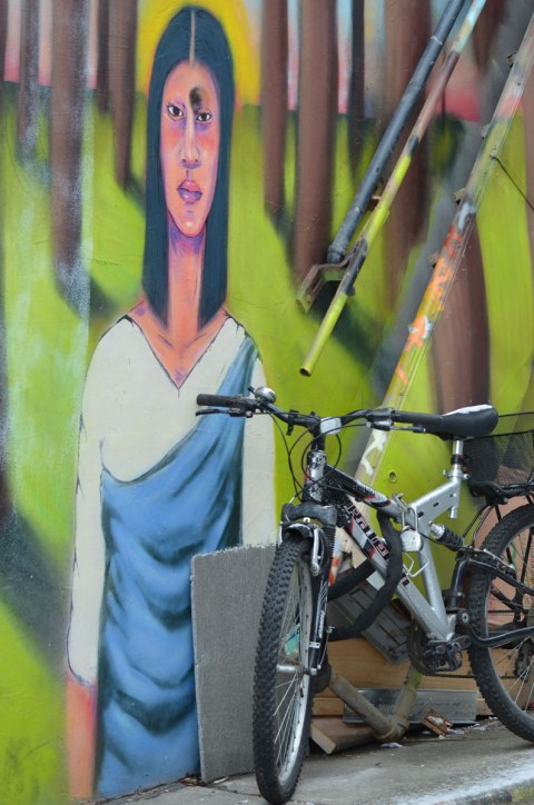 part of a mural in a Kensington lane of a woman with long dark hair and a white and blue dress, she is standing. In front of her is parked a bike.