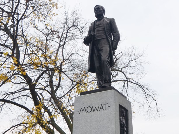 black statue of a man, Sir Oliver Mowat, standing with a book in one hand, and the other hand behind his back. The figure is on top of a grey stone rectangular column, autumn tree in the background. 