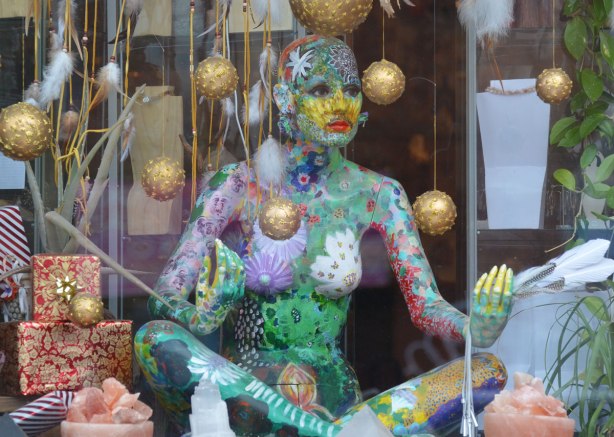 mannequin in a window sitting in a yoga position, and covered in painted patterns, multicoloured, gold Christmas balls are hanging from the ceiling around her.