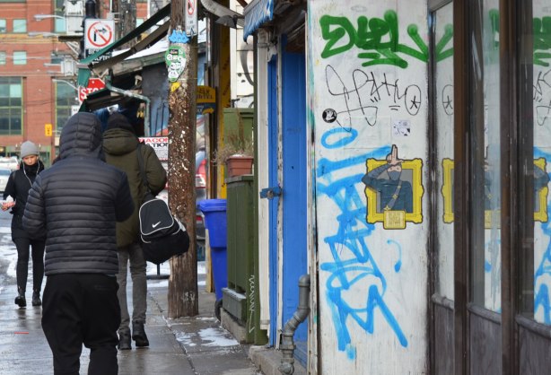 people walking in Kensington, walking past a wall with a pasteup on it of a man in uniform saluting he's drawn from the waist up and is in a yellow frame.