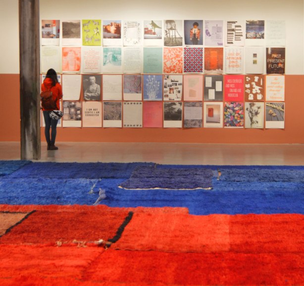 Part of an exhibit by Yto Barrada of a series of posters printed on paper and loosely tacked to the wall, of images and words. They are arranged in a grid of 12 x 8 pictures. A woman is standing in front of them, reading the words on one. Blue and red carpets are on the floor in the foreground. 