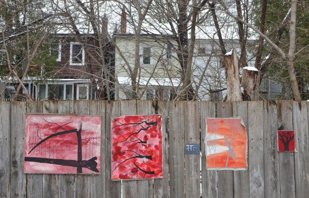 small amateur paintings displayed on a wood fence, with trees and houses in the background, snowy day, Craven Road in Toronto, two paintings of black trees (no leaves) on red, and one grey tree on orange background,