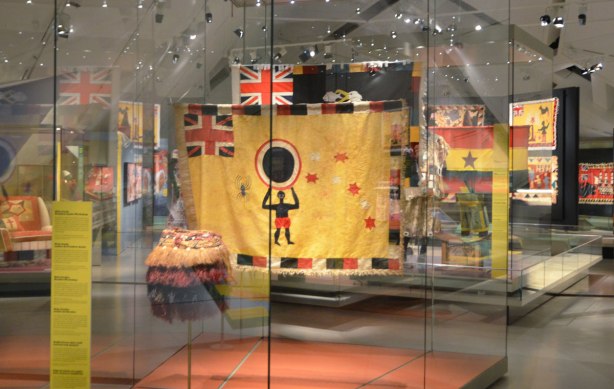 museum exhibit of asafo flags from Ghana, colourful flags of militia groups in yellows, reds and black. Many are hanging in display cases. 