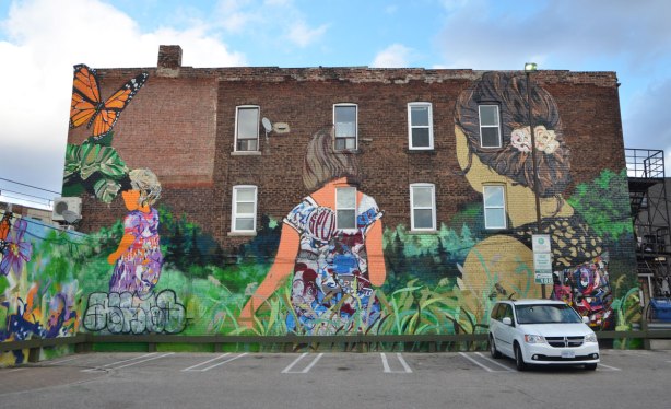 mural on the side of a brick building. Large. Three woman walking through long grass and chasing butterflies. A Stroll along Garrison Creek. Representing Emily Bickford and her daughters Beatrice and Grace. 