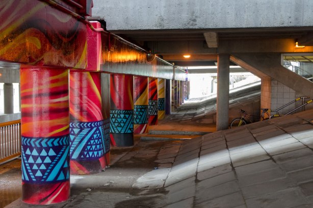 pillars and supports under an overpass that have been painted in bright colours by smoky and shalak - red pillars with blue geometric patterns in a band around it near the bottom 