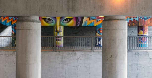 looking across Wilson Avenue, under the Allen Expressway towards a mural that has been painted on the pillars and supports on the other side. A face is painted there. 