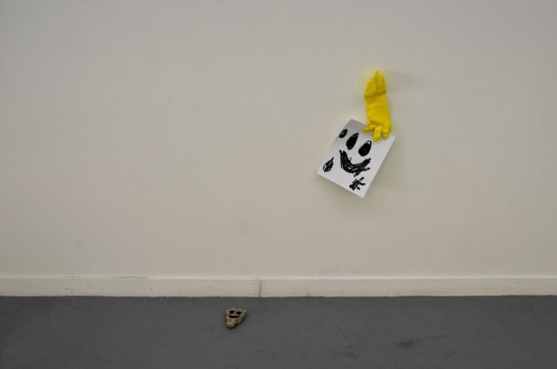 a black happy face scrawled on a piece of paper, a yellow rubber gloved taped to the wall that is holding the paper. A small rock on the floor with a happy face painted on it. 