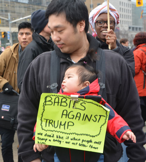 a man carries a baby in front of him, with a yellow sign that says Babies against trump, marching in the Womens March in Toronto with other men and women. 