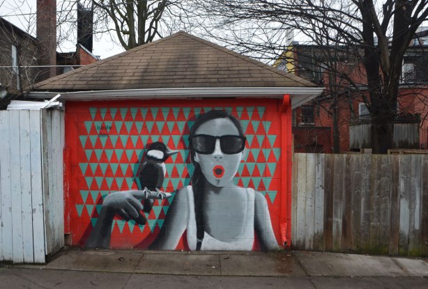 a birdo mural on a garage door of a woman in grey tones with a round red mouth in the shape of an O. she is holding her hand up near her face and a bird is sitting on it. The background of the mural is red and greenish triangles.