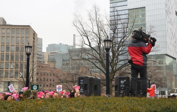 camera man stands on a high level of ground along with some large black speakers. The heads of some women can be seen , all wearing pink hats for the Womens March 