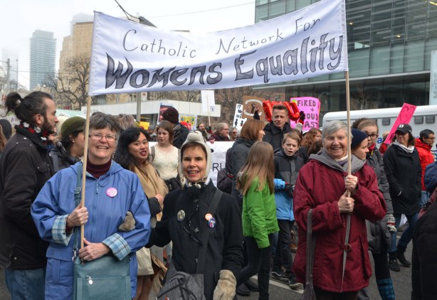 4 women pose for a picture, two are holding up a banner that says Catholic Network for WOmens Equality. Womens March, toronto . Lots of other eople, men, women and children, walking with them, 
