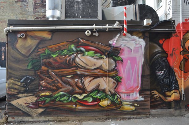 from a mural by elicser along the exterior wall of the dirty bird chicken and waffle restaurant - a man is sitting at a table in the restaurant . he has a very large chicken and waffle sandwich in front of him as well as a pink milkshake. The red and white straw of the miclkshake is actually a venting pipe 