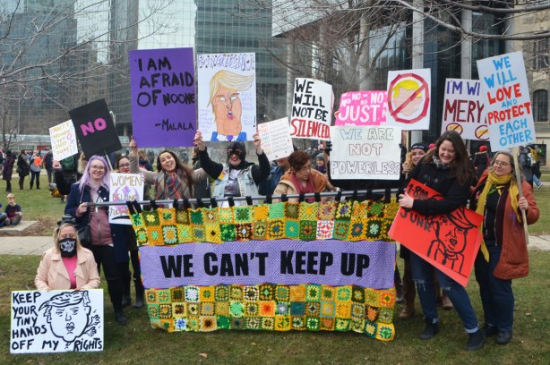 a group of women carrying a banner that has been made of many crocheted granny squares and the words we can't keep up. womens march in Toronto, waiting at Queens Park for the march to begin. 