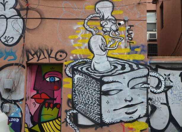 a street art painting on the back of a building in an alley, a surreal piece with a cube with one side as a face, two creatures are floating out of the top of the cube - they are tethered to each other. The bottom creature is half in the cube and the surface of the cube looks like liquid.