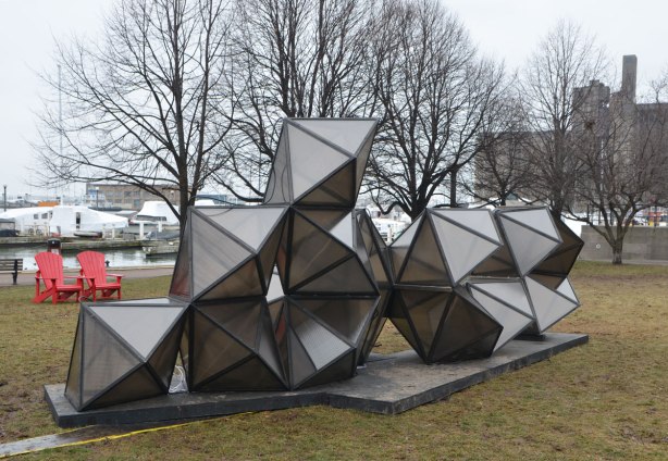 public art installation in a park along Lake Ontario, diamond shapes stacked in a pile, 10 pieces altogether, 5 on the bottom, 4 on the next row and one on top, grey in colour, bare trees and boats covered for the winter are in the background. 