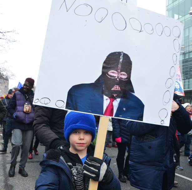 a young boy in a blue hat carries a placard that he's made that says Noooooo and has a picture of Donald Trump with a black Darth Vader mask on. 