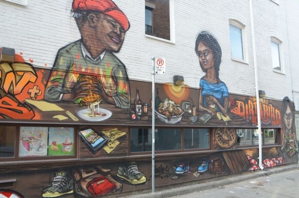 from a mural by elicser along the exterior wall of the dirty bird chicken and waffle restaurant - a woman and a man are sitting at a table in the restaurant eating chicken and waffle sandwiches