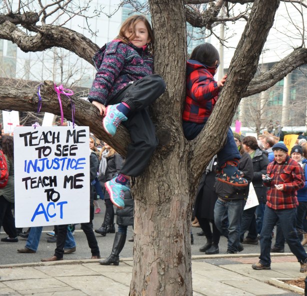 a young girl in a purple jacket is sitting on the branch of a tree. Her sign is strung over the branch and it says Teach me to see injustice teach me to act. People in the Womens March, toronto are walking past her in the background. 