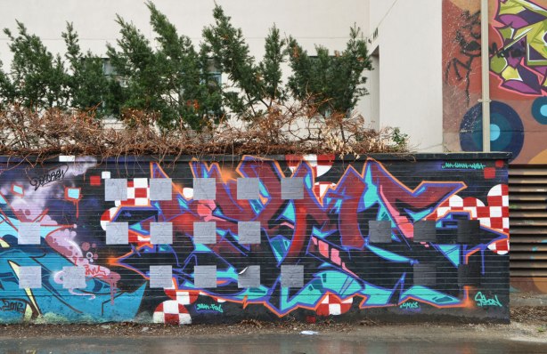 a grid of grey paper paste ups has been put on top of a abstract street art painting on a wall in an alley.